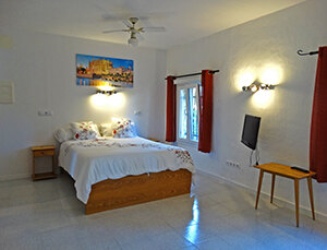 A guest room with white walls, a double bed and red curtains, a framed cove print above the headboard